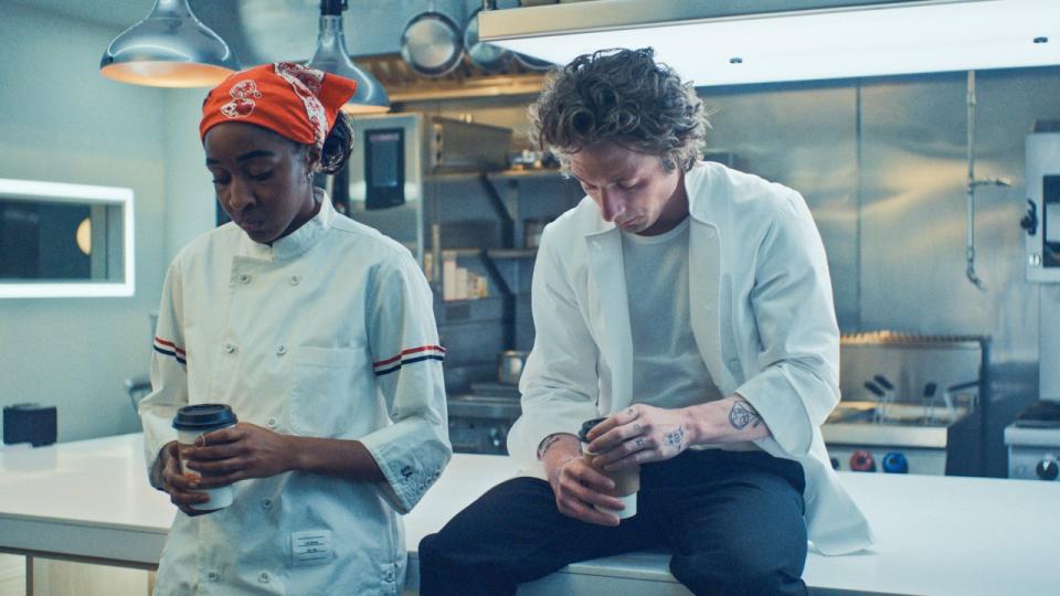 Ayo Edebiri standing and looking down while holding a cup and Jeremy Allen White sitting on a counter holding a cup looking down.