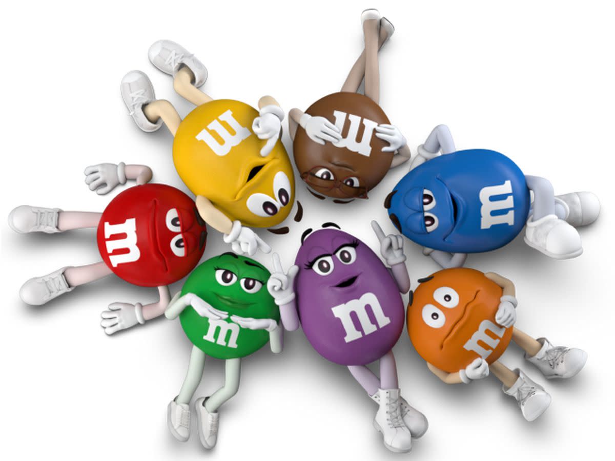 Top-view of updated six M&M's character laying on their backs in a circle, looking up at the camera, on a white background, 2023