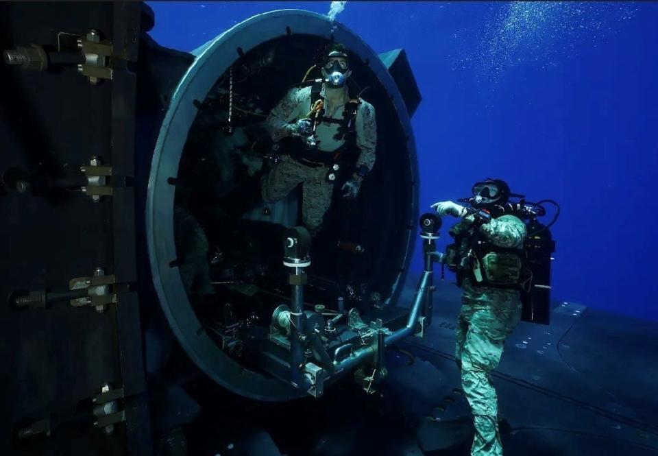 Two US Marines swim out of the dry dock shelter of a US Navy submarine in blue ocean water.