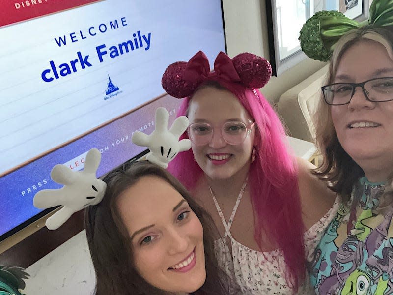 Selfie of the writer and family in front of a sign that says "Disney's Yacht Club Resort Welcome Clark Family." The writer wears a headband with Mickey Mouse hands, another girl with pink hair wears pink mouse ears, and the mother wears green mouse ears