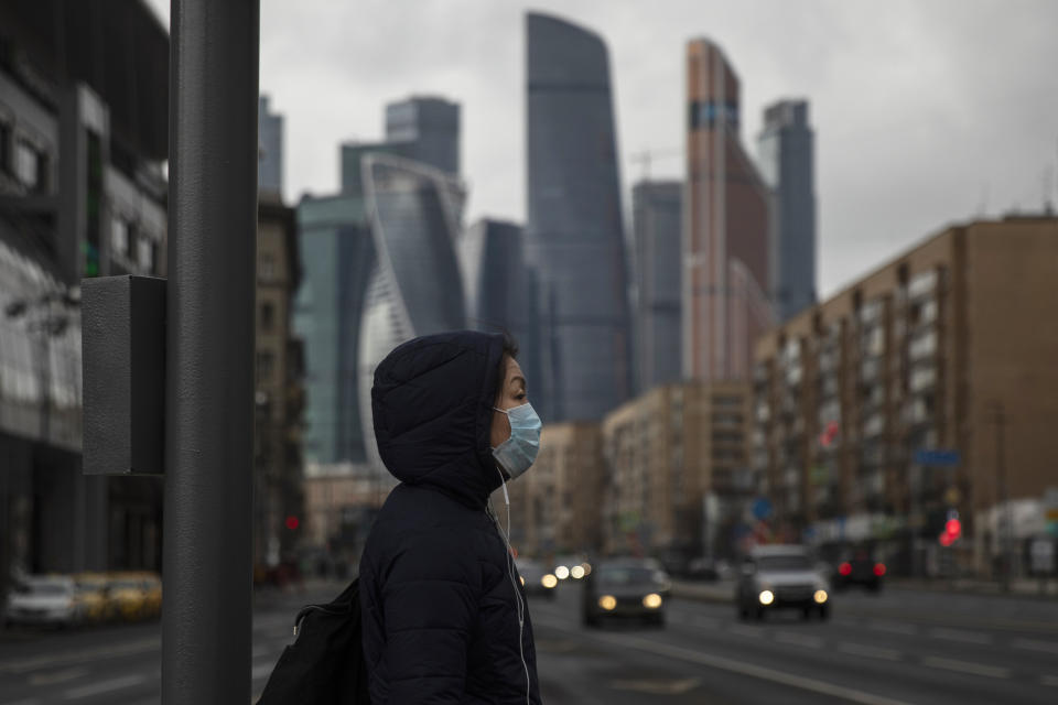 A woman wearing face mask waits to cross a nearly empty road due to residents taking the advice of staying at home to avoid the spread of the coronavirus, with Moscow City skyscrapers in the background, in Moscow, Russia, Thursday, April 2, 2020. Online shopping for food and other supplies has boomed in Moscow after the authorities put most residents on lockdown. The new coronavirus causes mild or moderate symptoms for most people, but for some, especially older adults and people with existing health problems, it can cause more severe illness or death. (AP Photo/Pavel Golovkin)