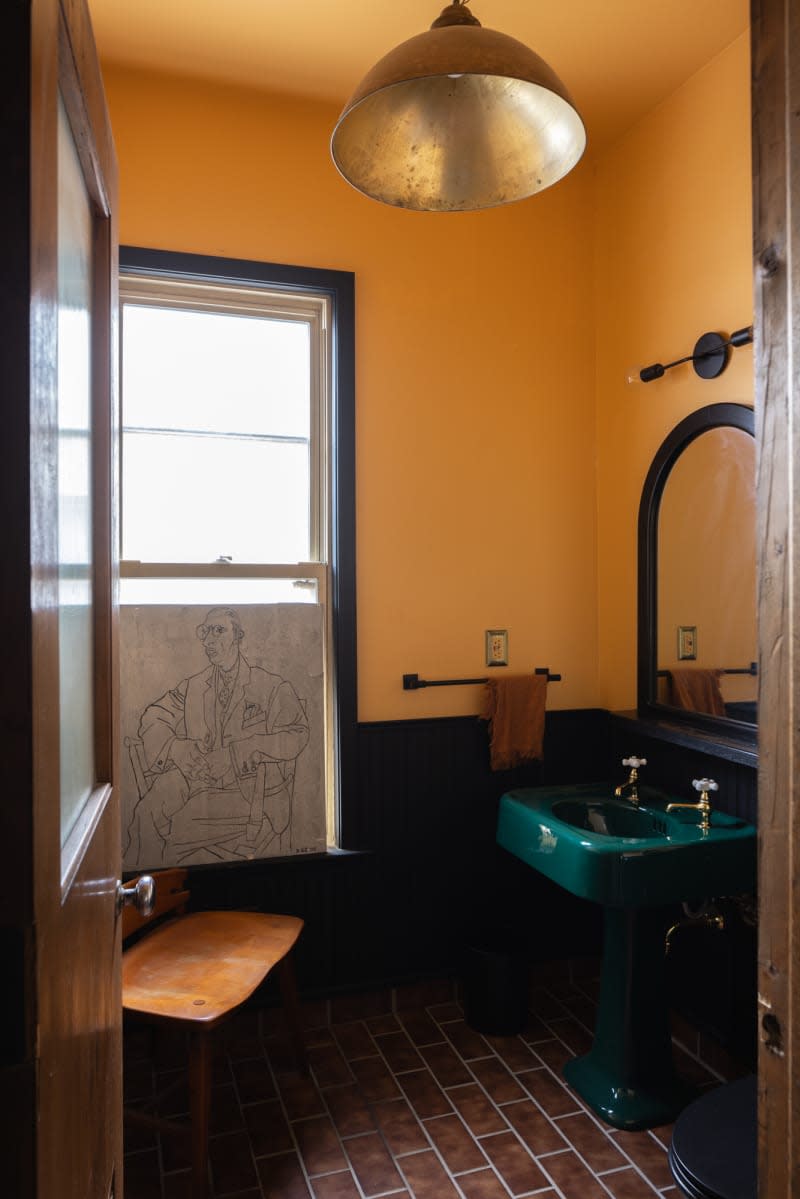 Green pedestal sink in gold bathroom with hanging metal pendant lamp and portrait art in window.
