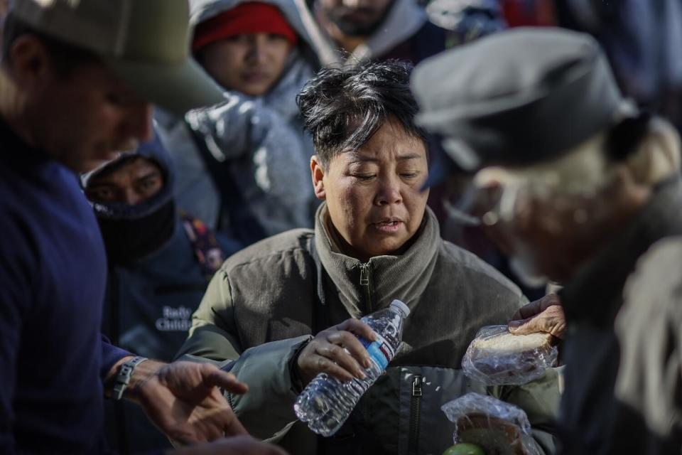 A woman in a crowd holding a water bottle a someone hands her a sandwich in a plastic baggie