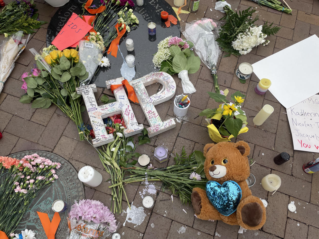 Flowers, candles and a teddy bear line a brick sidewalk with handwritten signs and large block letters HP.