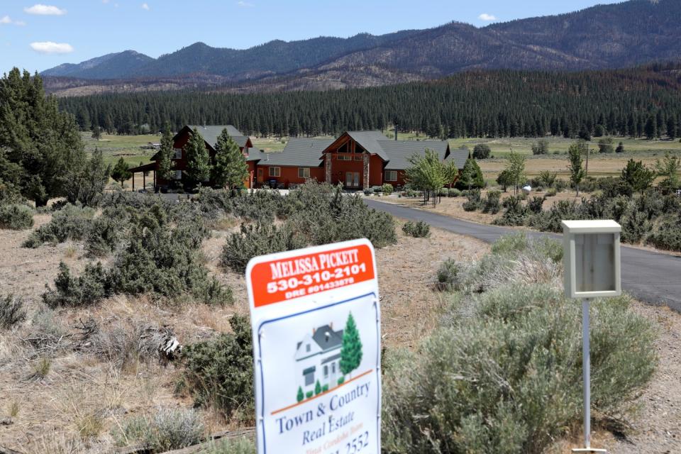 A "for sale" sign is posted at the end of a drive to a two-story house on open land with mountains in the distance.