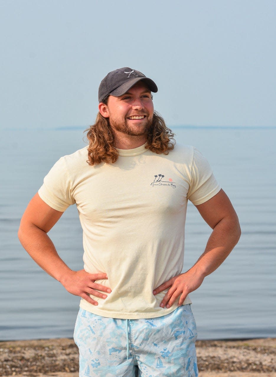 Alec Ochs stands in front of the Lake Erie shore in Port Clinton wearing one of his From Stream to Bay T-shirts. Each time a customer makes a purchase, Ochs picks up litter along waterways, most often at the edges of Lake Erie. Since launching the business in 2021, he has picked up nearly 1,600 pounds of litter.