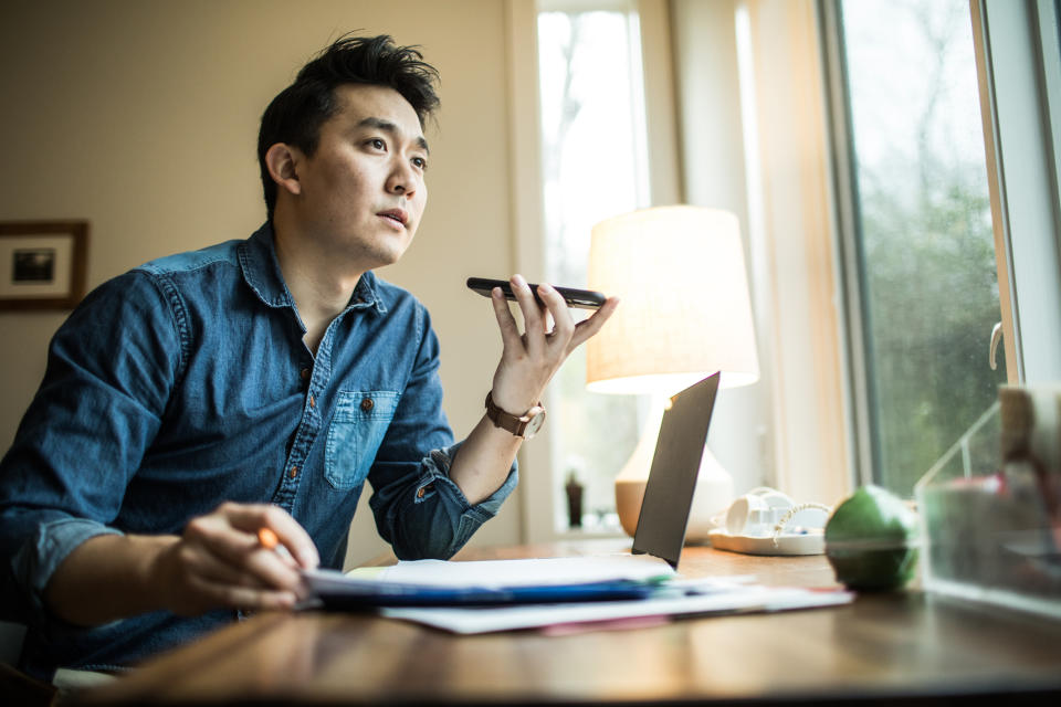 Man at a desk with a laptop speaks into a smartphone, possibly conducting business or reporting news