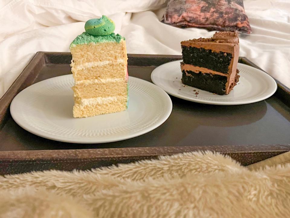 Slices of cake on white plates on a brown tray placed on top of white sheets and a fuzzy blanket. One cake is vanilla and has blue frosting and the other is chocolate