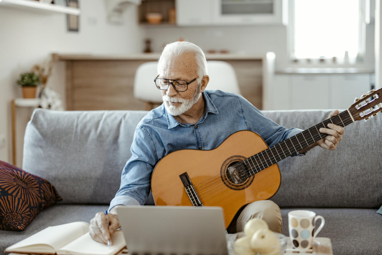 Bearded senior man making notes while watching online guitar course on laptop at home