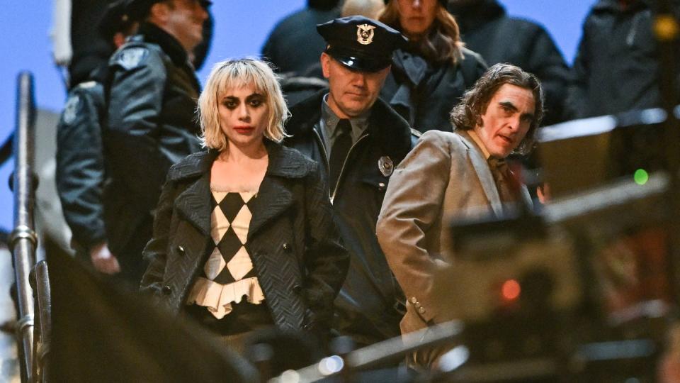 lady gaga and joaquin phoenix star at the camera while in character for the joker sequel, she wears a gray peacoat with a black and white diamond patterned shirt, he wears a brown suit, both have face paint on and a fake police officer stands behind them