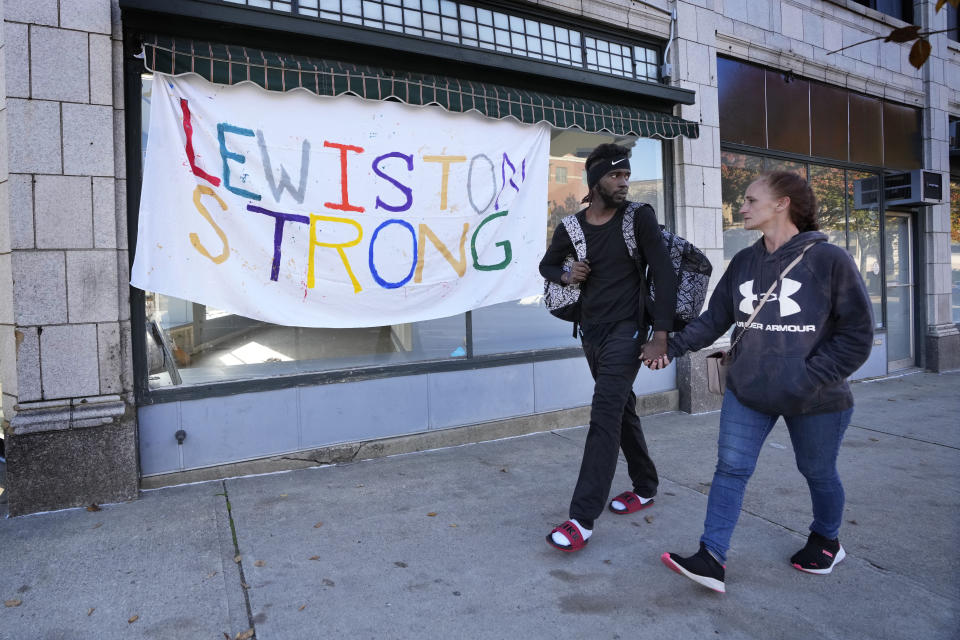 CORRECTS DATE TO OCT. 27, NOT OCT. 26 - A couple walks by a banner that was put up in response to this week's deadly mass shootings, Friday, Oct. 27, 2023, in Lewiston, Maine. Police are still searching for the suspect who killed at least 18 in separate shootings at a bowling alley and restaurant on Wednesday. (AP Photo/Robert F. Bukaty)