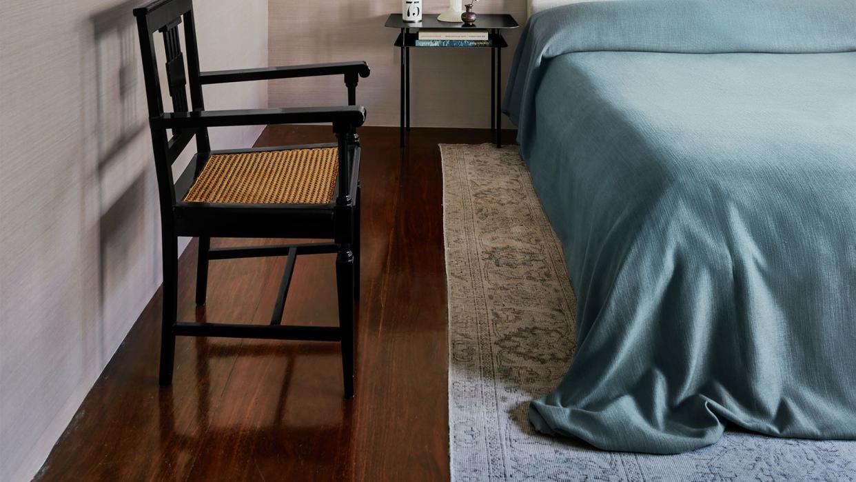 a bed with fabric headboard and a light blue linen cover, a black nightstand with a white lamp, a black lacquer wooden chair, a bleached kilim, and an old master painting and a woven metal artwork on walls