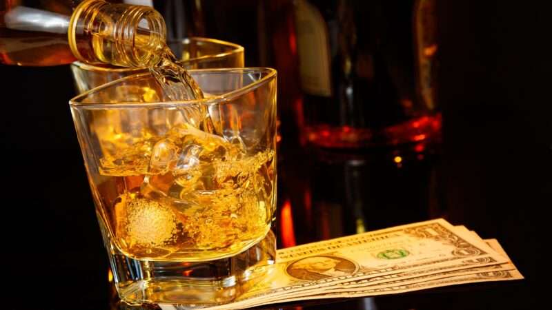 Whiskey being poured into a rocks glass over ice, with a few dollar bills on the bar underneath.