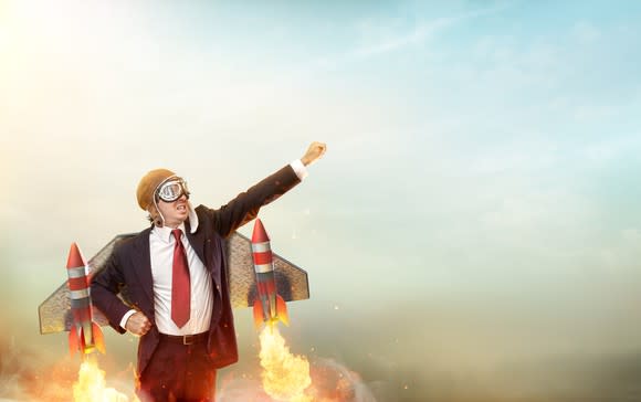 A man wearing a suit and a flight helmet and goggles raises his arm to the sky, as rockets strapped to wings on his back shoot out flames, suggesting an imminent launch.