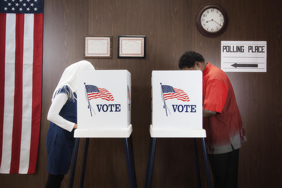 Two people casting votes in polling booths with American flags, "VOTE" signs, and "POLLING PLACE" sign visible on the wall. An American flag is in the background