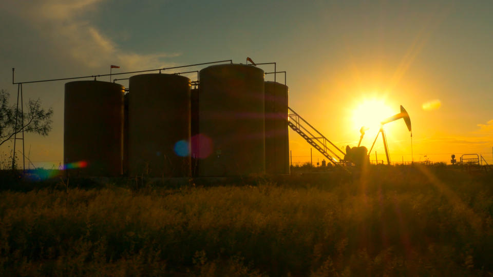 An oil pump and storage tanks with the sun rising in the background.