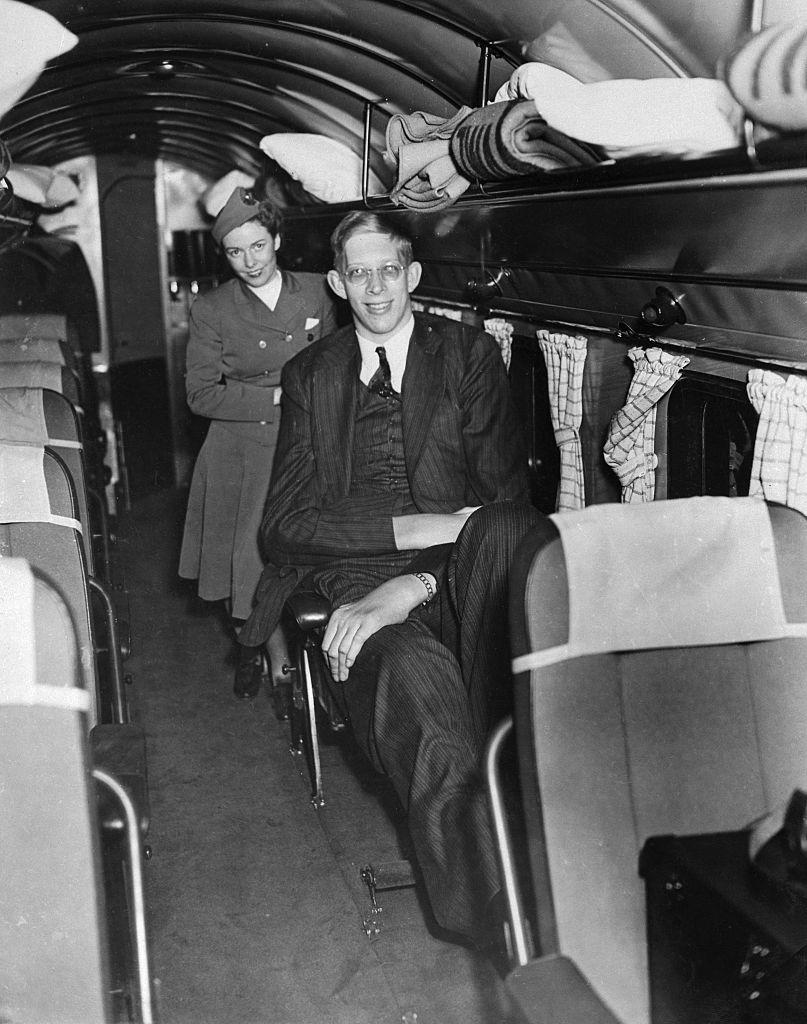 He's wearing a suit and sitting with one leg stretched out on an old, single-row plane and a flight attendant behind him