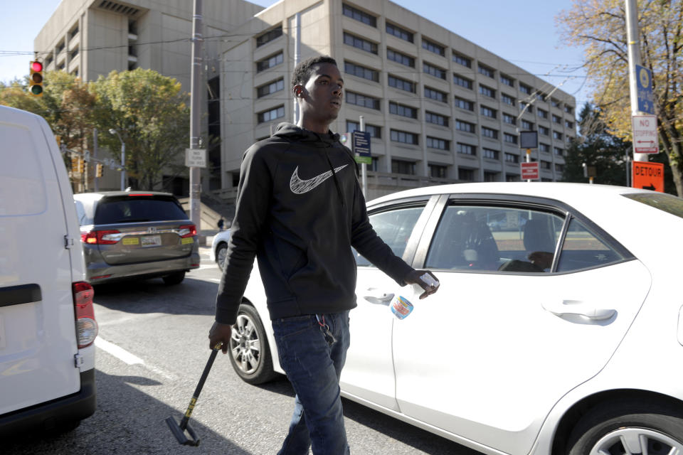 In a photo taken Thursday, Oct. 24, 2019, Nathaniel Silas walks through stopped traffic at a red light while looking for cars to squeegee in Baltimore. A debate over Baltimore's so-called squeegee kids is reaching a crescendo as the city grapples with issues of crime and poverty and a complicated history with race relations. Officials estimate 100 squeegee kids regularly work at intersections citywide, dashing into the street as red lights hit to clean windshields in exchange for cash from drivers. (AP Photo/Julio Cortez)