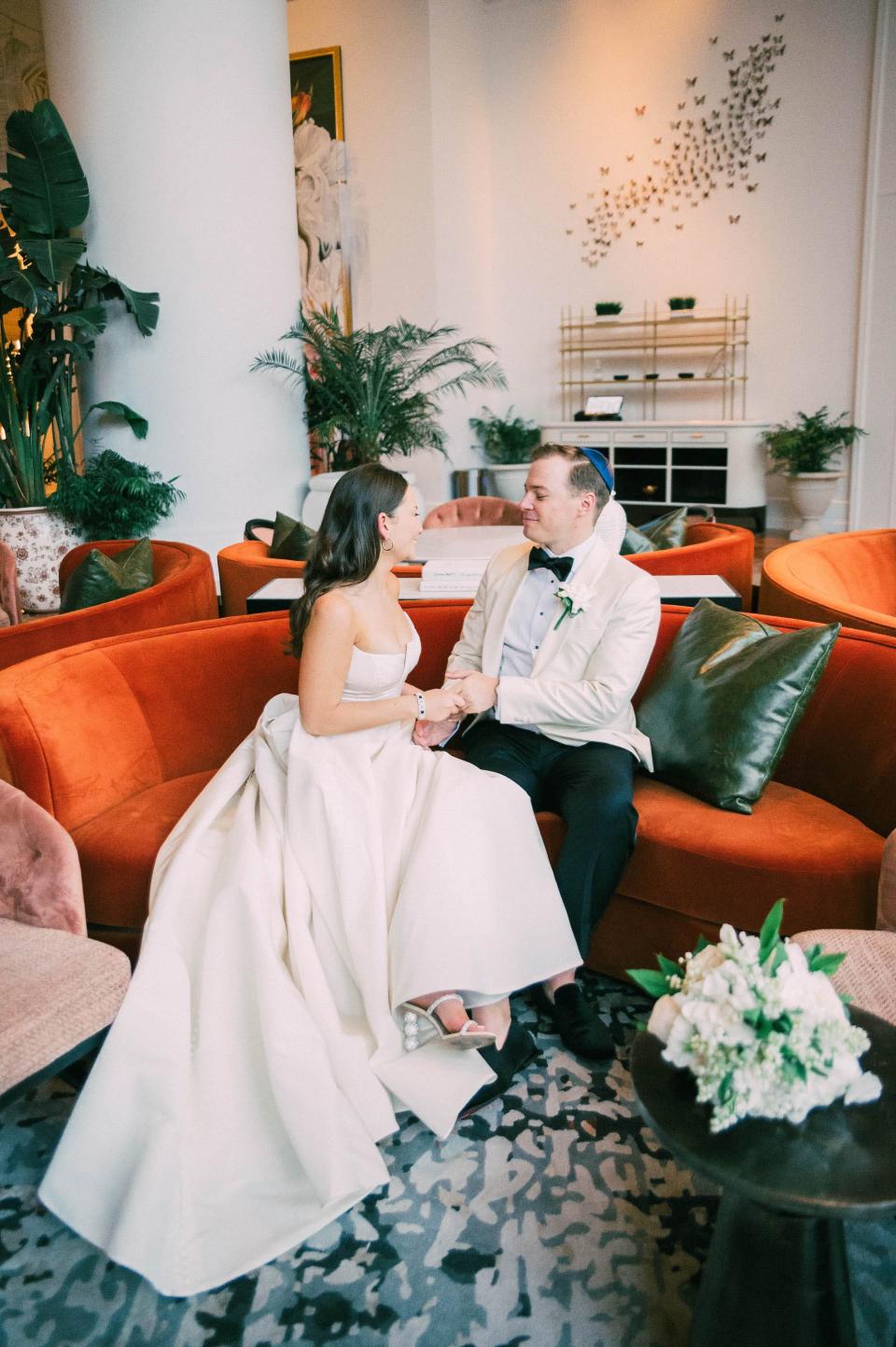 A bride and groom hold hands and look at each other on a couch.
