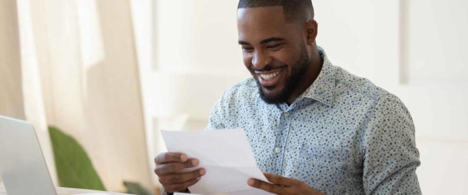 Smiling african American man sit at desk working at laptop reading good news in postal correspondence. He just got an unexpected tax refund.