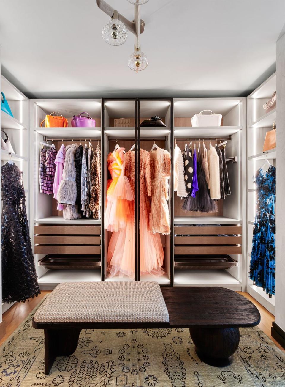 in the center of a walk in closet is a dark wood bench and a patterned rug, along the walls are sections with hanging clothing, drawers below, shelves, and storage cubbies with colored handbags above