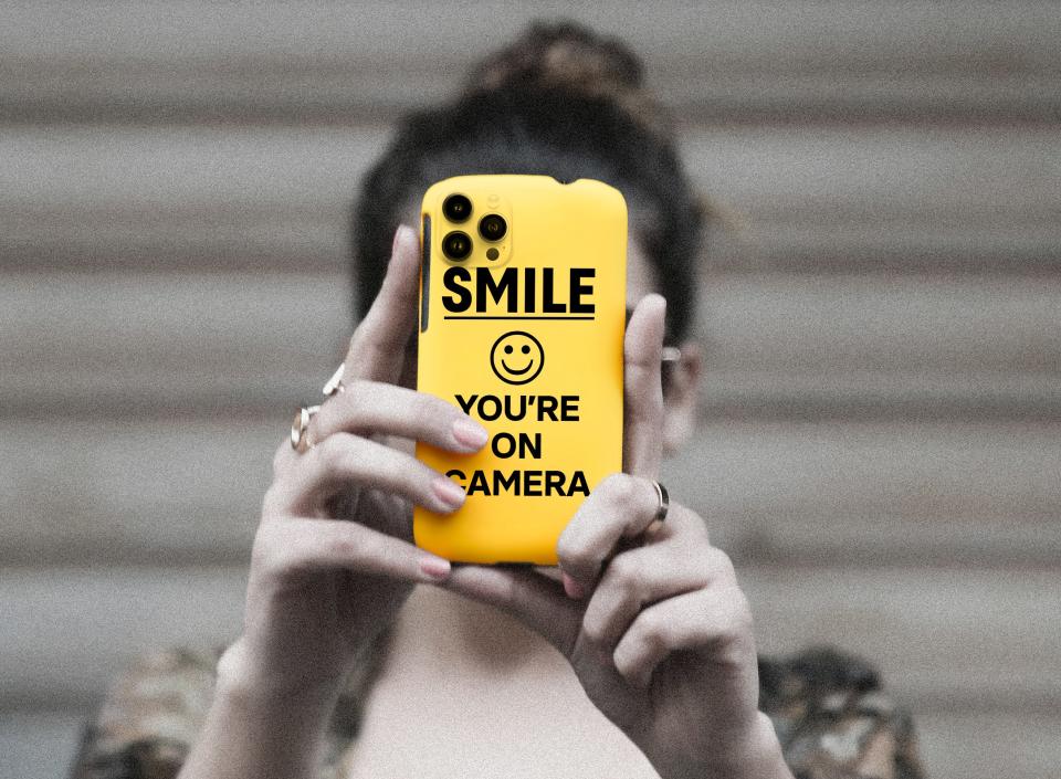 A woman holds a phone that resembles a sign that says "Smile, you're on camera"