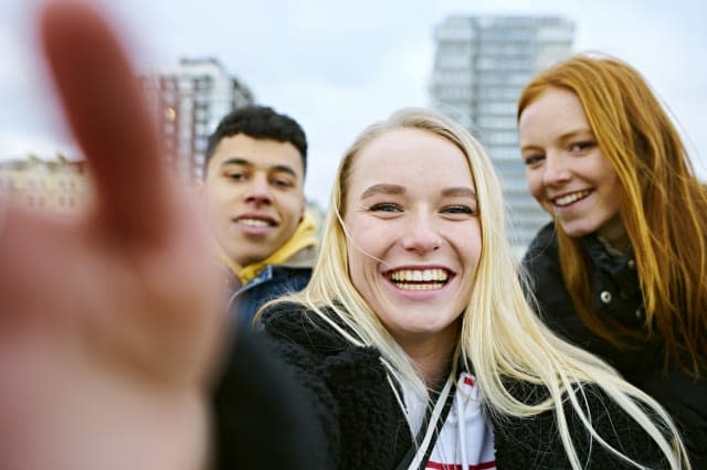 Three teenagers taking a selfie on their mobile phone taken from the phone's perspective with hand in view of the camera. Two friends lean in from both sides smiling at camera