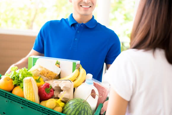 A smiling man delivers a tub full of groceries to a woman.