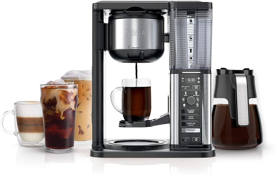 A coffee maker with a single glass cup underneath the dripping coffee. Variety of coffee drinks and a carafe are next to it.