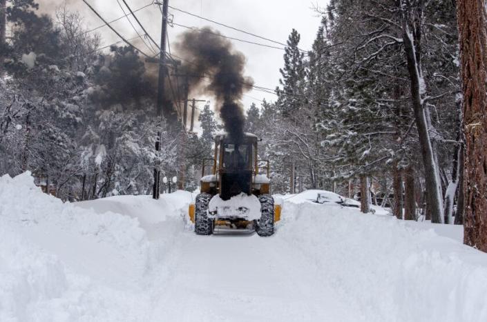 SUGARLOAF, CA - MARCH 01: A snowplow belches black smoke as the driver clears a path for vehicles past homes inundated with snow in the San Bernardino Mountains where successive storms dumped even more snow on Wednesday, March 1, 2023 in Sugarloaf, CA. (Brian van der Brug / Los Angeles Times)