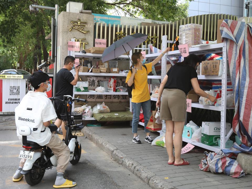 Metal shelves sit outside the fence of a gated community and four people search for their packages on the shelves.