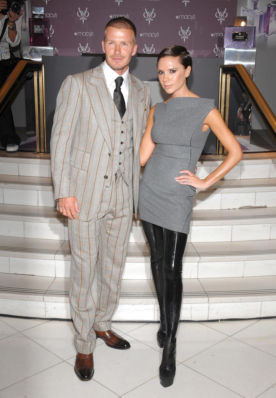 David Beckham and Victoria Beckham pose for a photo at the launch of the Beckham Signature Fragrance Collection at Macy's Herald Square on September 26, 2008, in New York City. David wears a gray, three-piece suit with pinstripes and a black tie, while Victoria wears a gray shift dress with black, thigh-high boots.