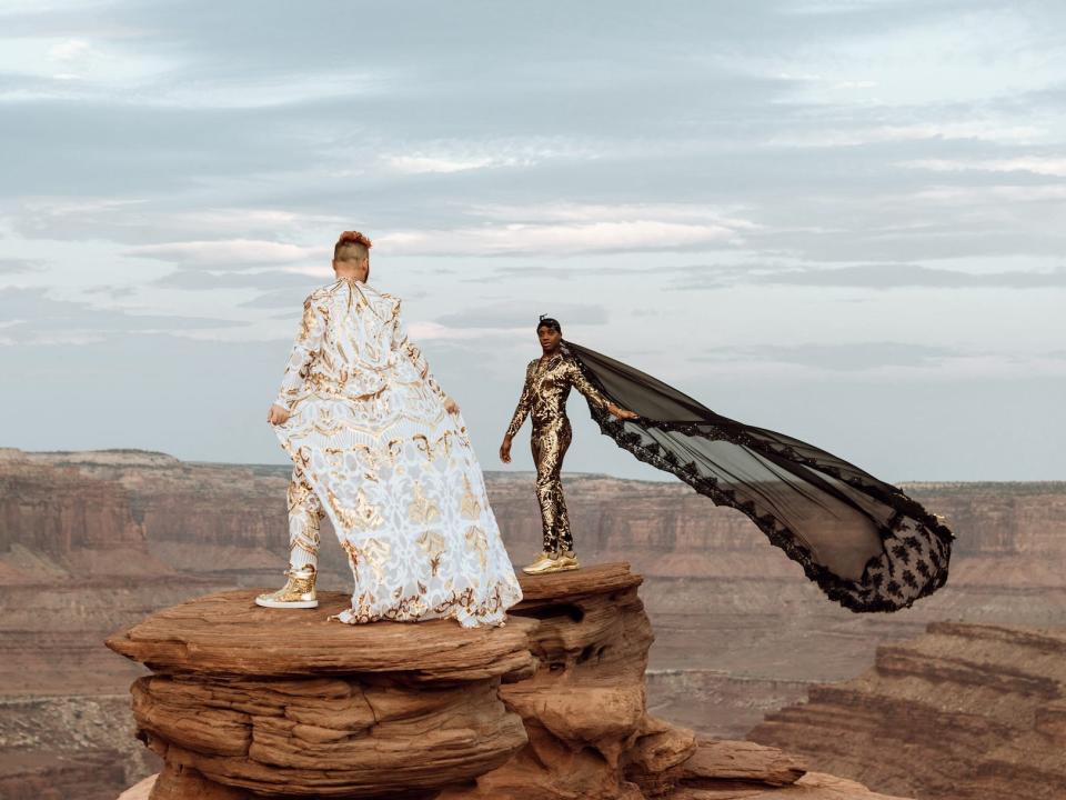 Two grooms stand in their wedding attire on a rock.