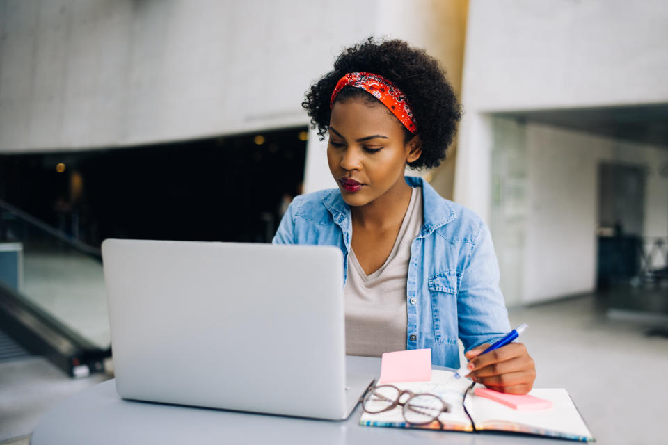 Young African female entrepreneur sitting at a table in a modern office building lobby working on a laptop and writing notes in her planner