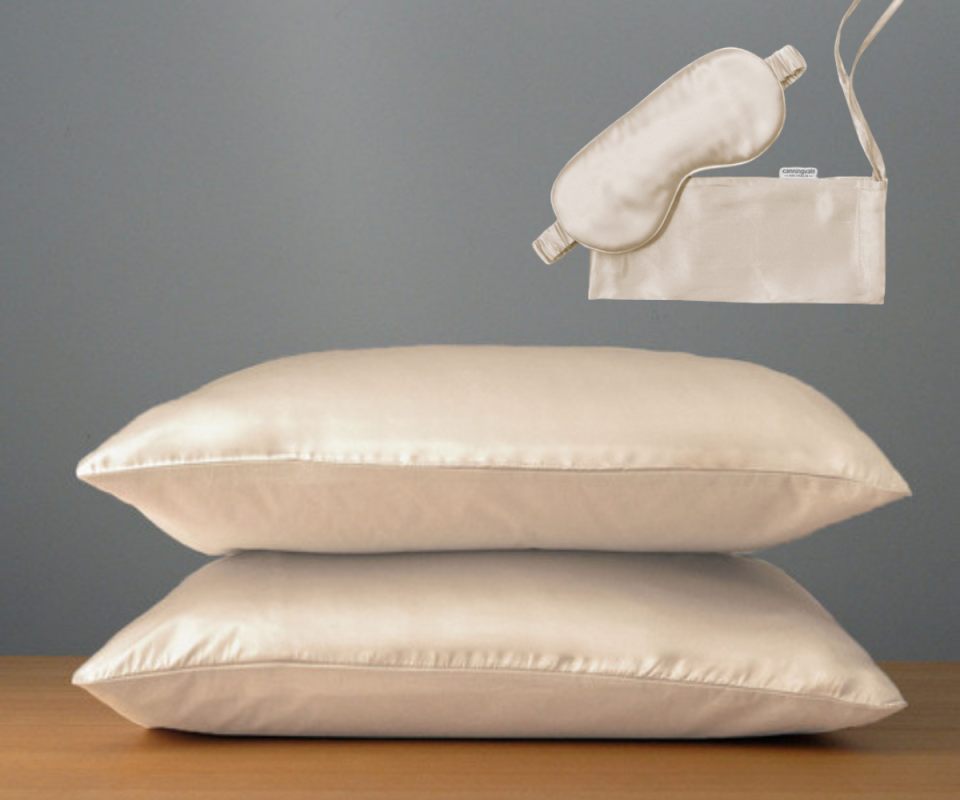 Two pillows in champagne silk pillow cases sit ontop of each other on a timber table against a grey background with matching champagne silk eye mask above.
