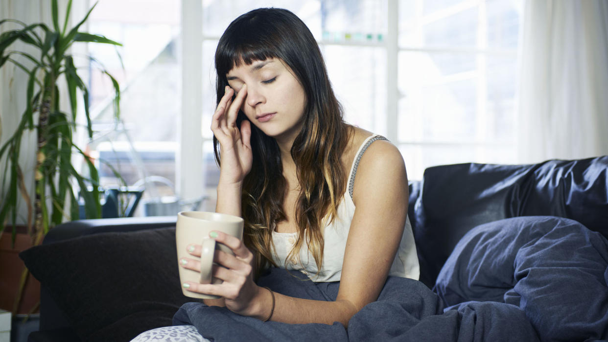  Woman sat up, rubbing her eyes and looking tired, with a mug in her hand 