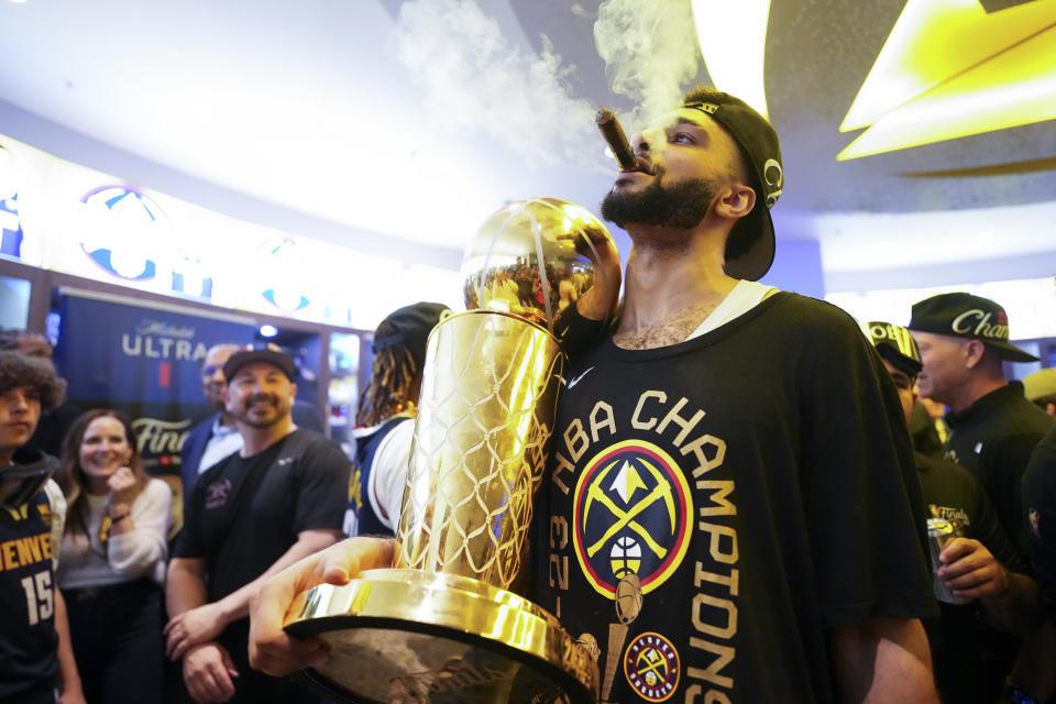 Denver Nuggets point guard Jamaal Murray smokes a cigar inside the locker room while holding the Larry O'Brien Championship Trophy after winning the NBA Championship against the Miami Heat in Game 5 of the NBA Finals.  (AP Photo/Jack Dempsey)
