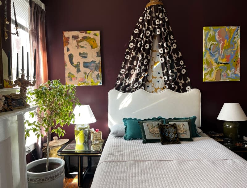 Marimekko fabric draped above white bed with white bedding in eggplant bedroom with colorful artwork and potted plant.