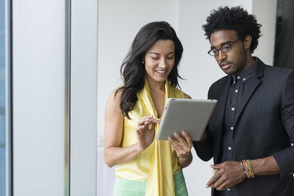 Two individuals stand by a window. A woman in a sleeveless blouse is holding and pointing at a tablet, while a man in glasses and a blazer observes