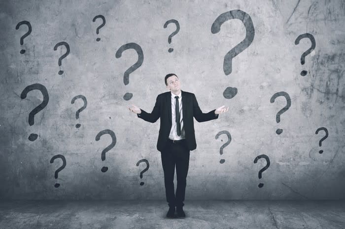 Businessman shrugging and holding hands out with question marks on the wall behind him