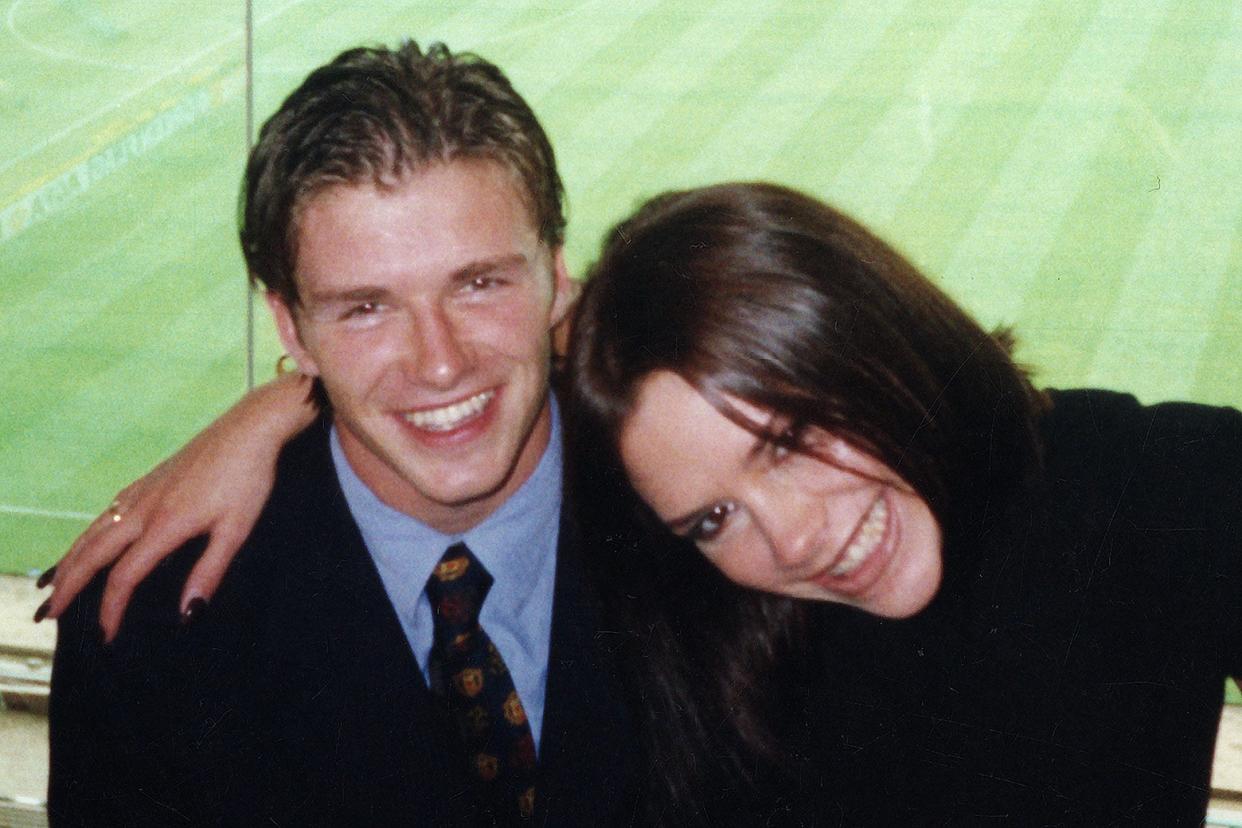 A younger David and Victoria Beckham, in their twenties, smiling with their arms around each other during a football match.