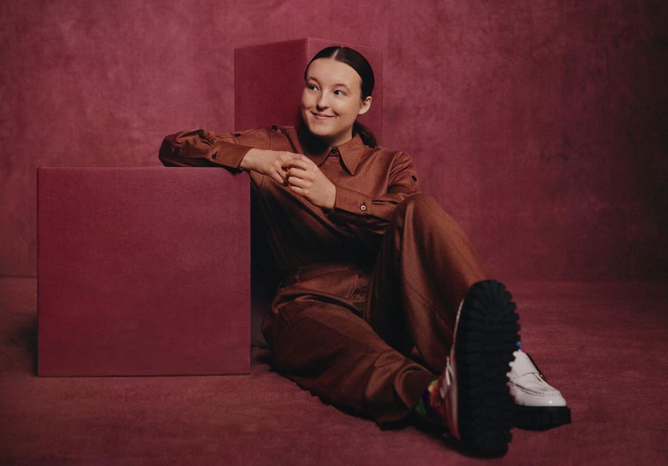 Actor Bella Ramsey with an impish smile, sitting before a burgundy backdrop for a portrait.