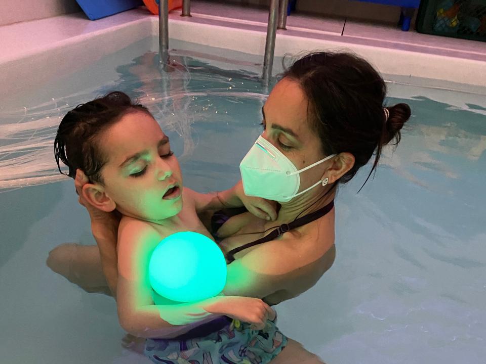 Mom Deborah Vauclare, who is wearing a black swimsuit and a face mask, holds her son, Leo, wearing blue swimming shorts, during an aqua and light therapy session inside a pool.