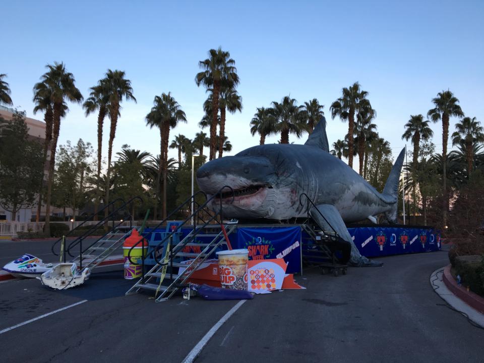 A convention float that has a huge megalodon on top of it on the side of a road with palm trees in the distance.