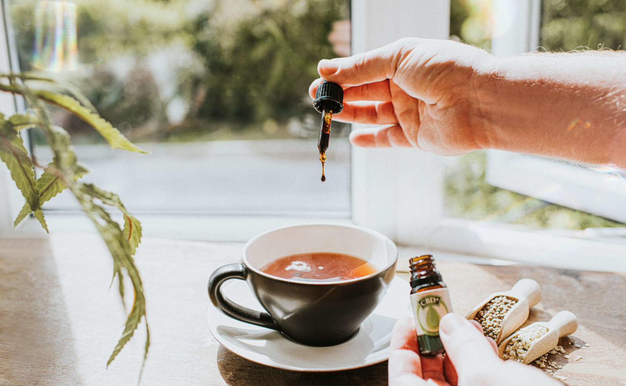 Lifestyle image of a hand holding a dropper filled with CBD oil and dropping it into a hot cup of tea sitting on a saucer on a table surrounded by Cannabis leaves in a sunny interior. Other hand holds Bottle of CBD oil closer to the camera, crushed and whole hemp leaves in scoops also sit on the table. Space for copy.