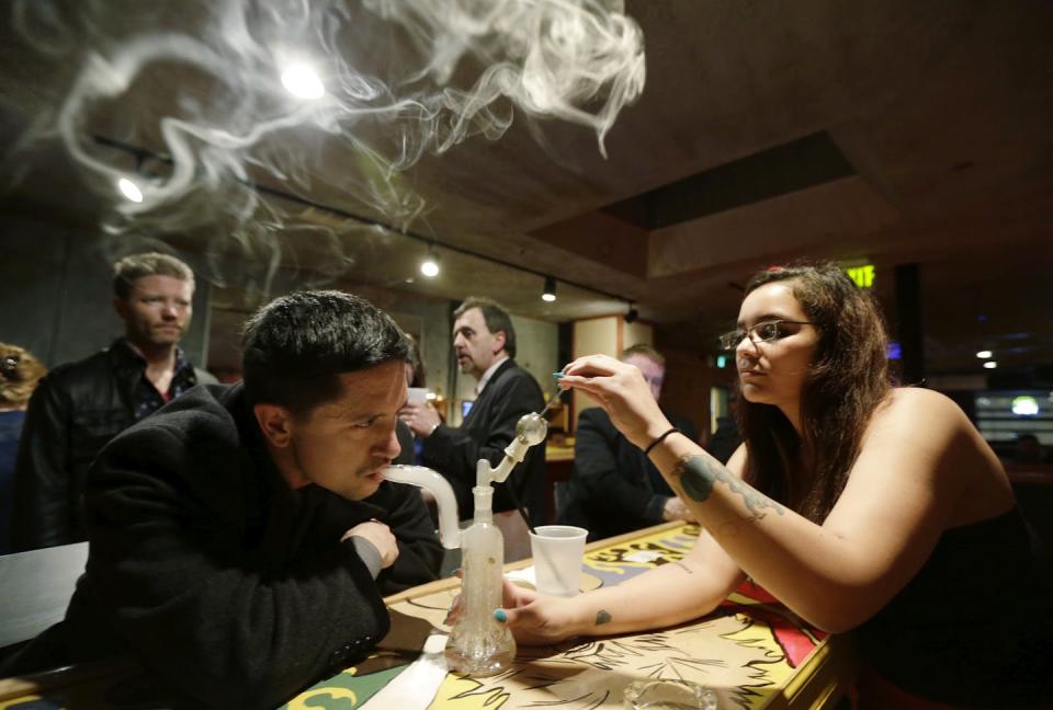 Un hombre inhala humo de marihuana de una cachimba de cristal en una habitación con otras personas alrededor.