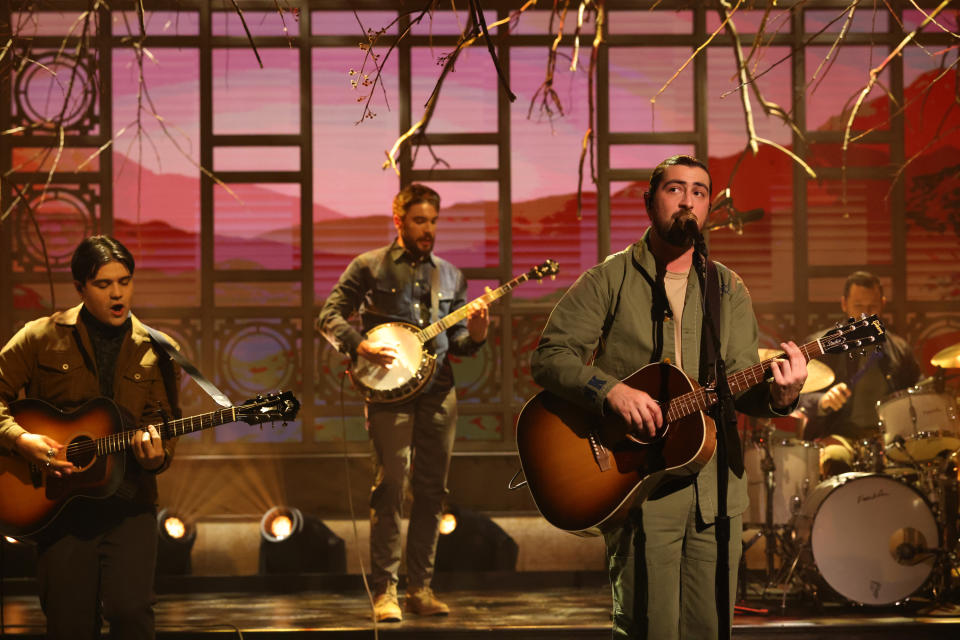 Three musicians performing, one with a banjo, one singing and playing guitar, and a drummer in the background