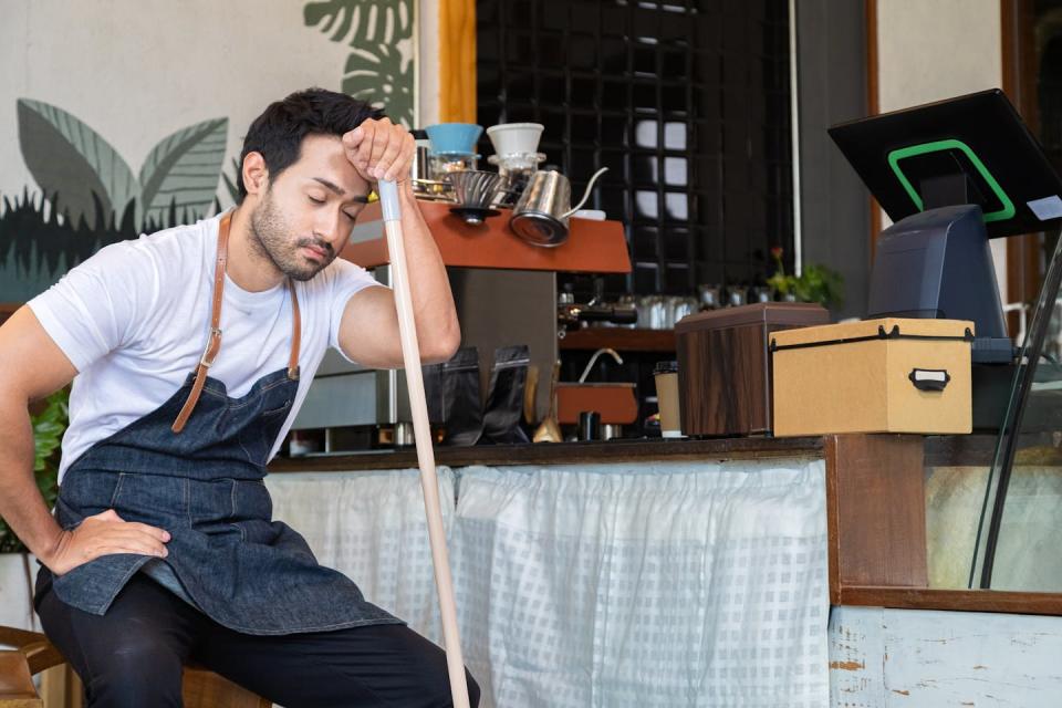 Man wearing an apron, sitting down resting his forehead on the end of a broom, looking tired.