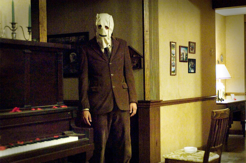 A person stands in a dimly lit room wearing a suit and a creepy mask. A piano and framed pictures are in the background