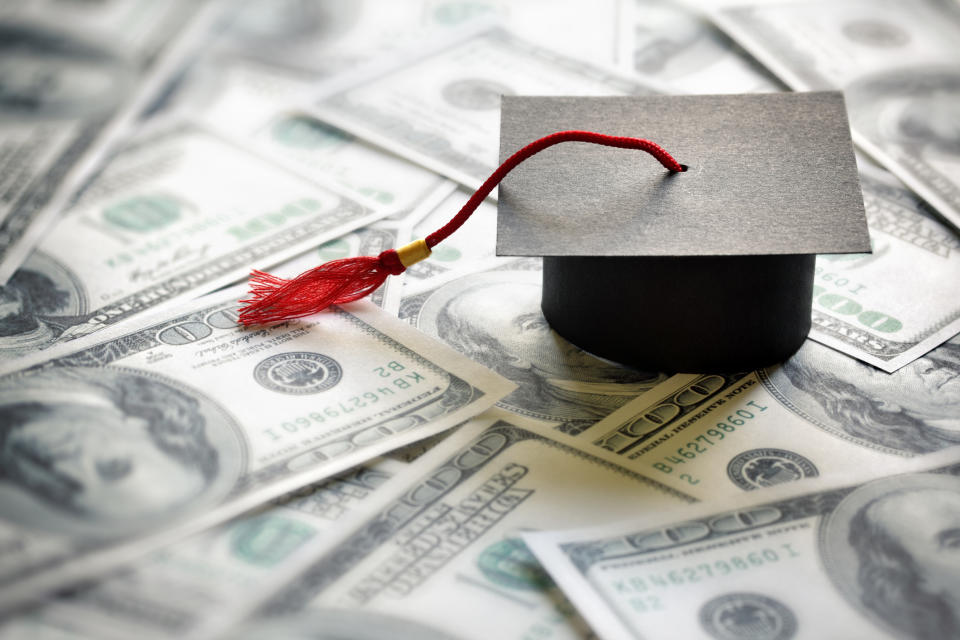 A miniature graduation cap sits atop a scattered pile of U.S. hundred dollar bills.